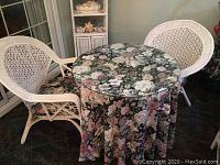 Set of round iron table and two white wicker chairs with floral fabric skirt and glass top in place.