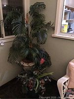 Full view of the tall artificial palm tree with large green leaves and pink flower in a corner next to a window, showing the decorative planter and green pot below with an additional artificial plant.