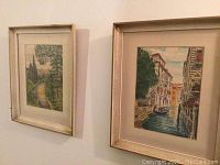 Both framed paintings displayed on a wall, showing Italian canal and nature scenes, with light-colored frames.