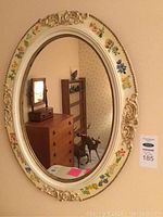 Front view of oval framed mirror showing full frame and reflection of room interior including chest of drawers and decorative animal sculpture.