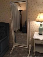 Full length mirror on white stand positioned in living room with upholstered furniture and lamp visible