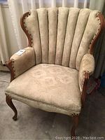 Front view of cream upholstered antique armchair with high scalloped back and carved wooden frame with nailhead trim at arms and seat.