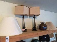 Two black table lamps with beige rectangular shades, additional white and cream pleated lamp shades, displayed on wooden shelf.