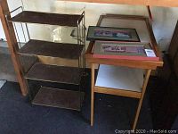 Full view of the vintage stand with three wood shelves next to a wooden side table with the two framed signs placed on top and leaning against the back wall.