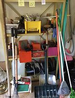 Wide view of metal shelving unit holding assorted garden tools such as rakes and snow shovels, various plastic storage boxes, and tool rack