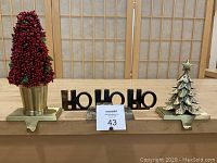 Front view of three stocking hangers: red berry tree, HoHoHo silver plated text, silver Christmas tree with star topper.