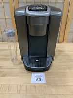 Front view of Keurig K-Elite K-90 coffee maker with clear plastic water reservoir on the left side and a drip tray at the base.