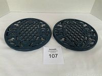 Two dark blue enameled cast iron round trivets on white background with lot number card between them
