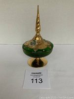 Front view of vintage green glass covered bowl with gold designs and tall finial lid on gold pedestal base.