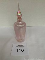 Full view of the pink iridescent glass perfume bottle showing the white swirl design and the stopper. Bottle stands on a flat surface with lot number card visible.