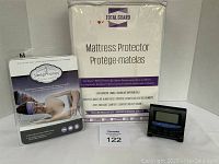 Photo showing Total Guard queen size mattress protector in packaging, SleepPhones sleep headphones box with imagery, and digital travel alarm clock with user manual.