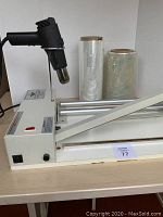 Front and side view of tabletop shrink wrap machine with black heat gun mounted, two rolls of plastic wrap behind it, and a metal arm for securing items in place during wrapping.