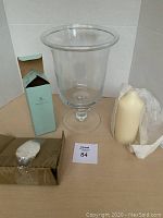 Photo of the glass hurricane lamp, pillar candle, and taper candle insert in packaging, displayed on a neutral background with lot number card.
