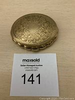 Closed top view showing detailed etched floral pattern on gold-tone lid with scalloped edge.