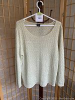Front view of the light green knit sweater hanging on a white hanger, showing its scoop neckline and long sleeves against a wooden screen background.