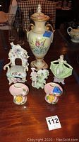 Photo shows six porcelain and ceramic decorative items on wooden surface including a detailed urn with lid, three figurines depicting cat on a chair, dress, and man with hat, and two small pink floral vases.