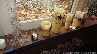 Four cream-colored candles with black star and ribbon designs on a wooden tray, including one candle wrapped with twine and a small glass votive holder with a Christmas tree silhouette.