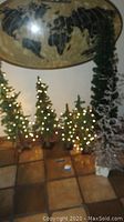 Group of 7 faux Christmas trees of various heights, some with white string lights turned on, standing on a tile floor under wall art.