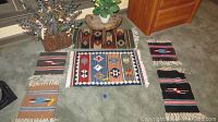 Six southwestern style woven rugs displayed on carpet, featuring geometric designs with fringe, along with a small wood stool and faux flower planter to the side.