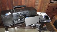 Sony CFD-550 boombox with detachable speakers, Bose Sound Dock portable speaker, Omron digital blood pressure cuff with manual, CDs and rack on glass top table in wood-paneled room.