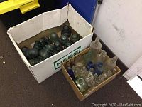 Two cardboard boxes containing multiple vintage glass electrical insulators and assorted glass bottles of varying sizes and colors including blue, green, and clear glass.