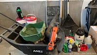 Wide shot showing Ames black wheelbarrow, Scotts turf builder seeder, Black & Decker orange and black lithium ion sweeper/vac, several garden tools and chemicals in the back.