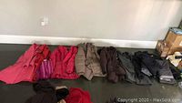 Wide view showing 7 coats and 2 ski pants laid on floor in a row in various colors