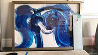 Wall art with large blue swirl design in stainless steel frame and rectangular gray glass clock leaning against wall.