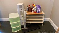 Photo showing shelving rack, plastic storage cabinet with three drawers, wooden turntable, and multiple cleaning supplies on top of shelves.