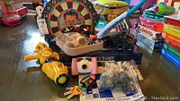 Photo of assorted toys and games including dartboard, Pictionary box, chess set, NERF bow and arrow, Fisher-Price camera and other toys.