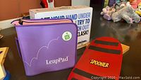 LeapPad purple carrying case with text and Klask game box in background on table with plush toys