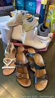 Photo showing white ice skates with pink blade covers, gray sneakers, white strap sandals, and black buckle sandals arranged on a table.