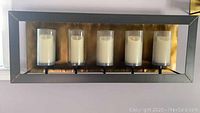 Front view of the metal framed candle holder mounted on wall with five cream-colored flameless candles inside cylindrical glass shades.