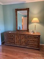 Front angled view of large oak dresser with attached mirror on a hardwood floor against painted wall, showing drawer and door layout, wood grain, and the lamp and glass decor on top.