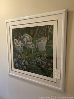 Framed print of a garden scene showing three white wicker chairs with cushions, small table, and various flowers in a green garden setting, with white frame and double matting.