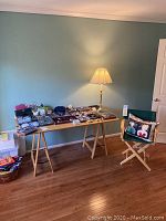 Full view of the sawhorse table with various items on top, the green director's chair with the pillow on it, set in a room with wooden floor and blue wall.