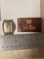 Close-up photo of the Lord Elgin gold filled wristwatch next to a ruler and the Lord Elgin certificate case