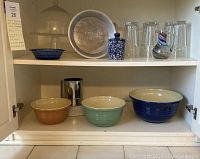 Set of three multi-colored Over & Back nesting bowls on bottom shelf showing colors brown, pale green, and dark blue; above clear drinking glasses, pie pan, ceramic canister, glass cake stand with dome, and blue glass dish.
