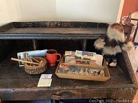Photo showing a dark rustic wooden desk with a wooden tray containing vintage postcards, a red ceramic mug, a round wooden dish with a mallet, and an Inuit-style child's doll standing beside the tray.
