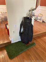 Full view of the black golf bag with clubs inside, standing on a patch of astroturf shaped like Oklahoma on a wood floor by a white wall.