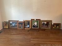 Photo of seven framed paintings arranged on floor against wall showing various rustic wooden frames and different subject matter paintings including fruit still life, portrait, wildlife, and nature