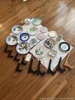 Photo of 19 hand painted porcelain plates, a white ceramic mug, a lidded porcelain box, and 8 plate display stands (7 black plastic and 1 wooden) arranged on a white cloth on wooden floor.