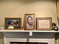 Display of three framed religious artworks on mantel including nativity scene, Jesus portrait, and Proverbs scripture; ceramic change jars labeled 'Hugs & Kisses' and 'Keep The Change' on either side.