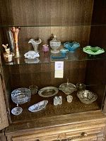 Full display cabinet showing both upper and lower shelves holding assorted vintage decorative glass pieces mostly hobnail glass and carnival glass with iridescent finish.