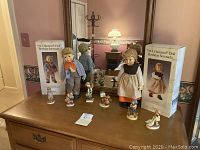 Photo showing 6 M.I. Hummel dolls and figurines on a wooden table in front of a mirror, including 2 large dolls and 4 smaller figurines.