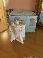 Photo of Lladro figurine of three angels reading a book, with original box behind.