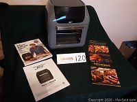 Photo of Air Fryer Pro unit with owner's manual, Eric Theiss cookbook, and three additional cooking guides on a green fabric background.
