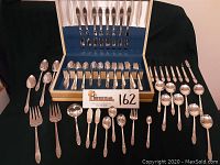 Full view of all flatware laid out in front of the open storage chest showing large assortment of forks, spoons, and knives with intricate floral pattern handles.