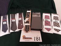 Photo showing a group of ten ties from various brands, all wrapped in original packaging, placed on dark green background with lot number card.