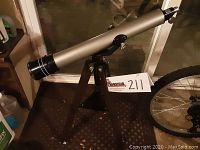 Left side view of silver telescope tube mounted on dark wooden tripod next to bike wheel and other items.